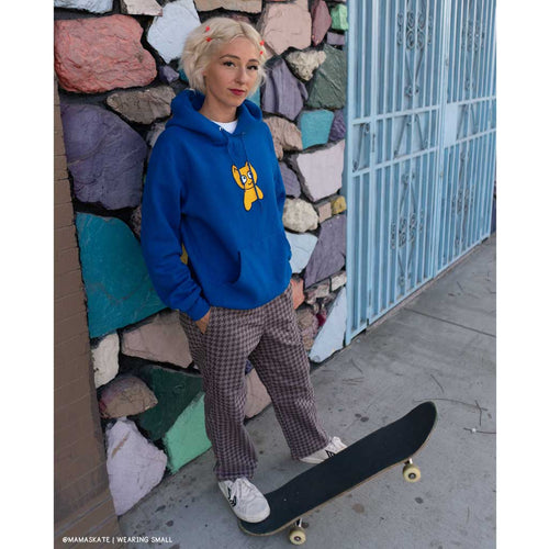 Person wearing a blue hoodie with a yellow graphic, plaid pants, and white sneakers standing next to a colorful stone wall.